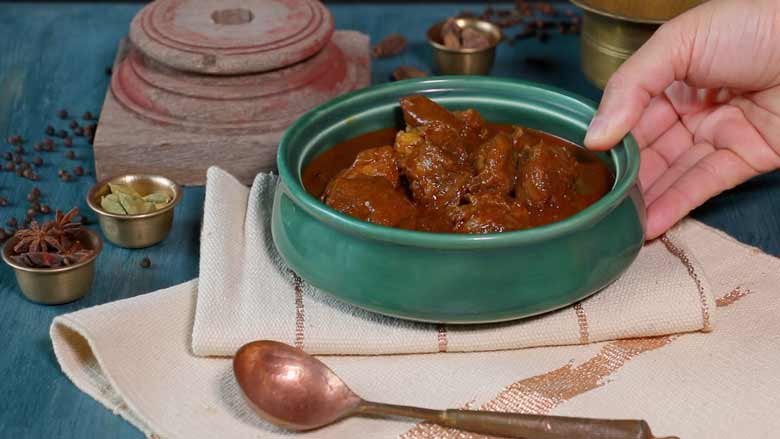 A hand is seen placing a emerald green ceramic bowl full of Rampuri Mutton Korma onto a striped cream and gold woven table mat with a copper ladle in the foreground and small brass bowls with whole spices and an antique pink wooden piece in the background on a blue painted wooden table - Taar Gosht Recipe